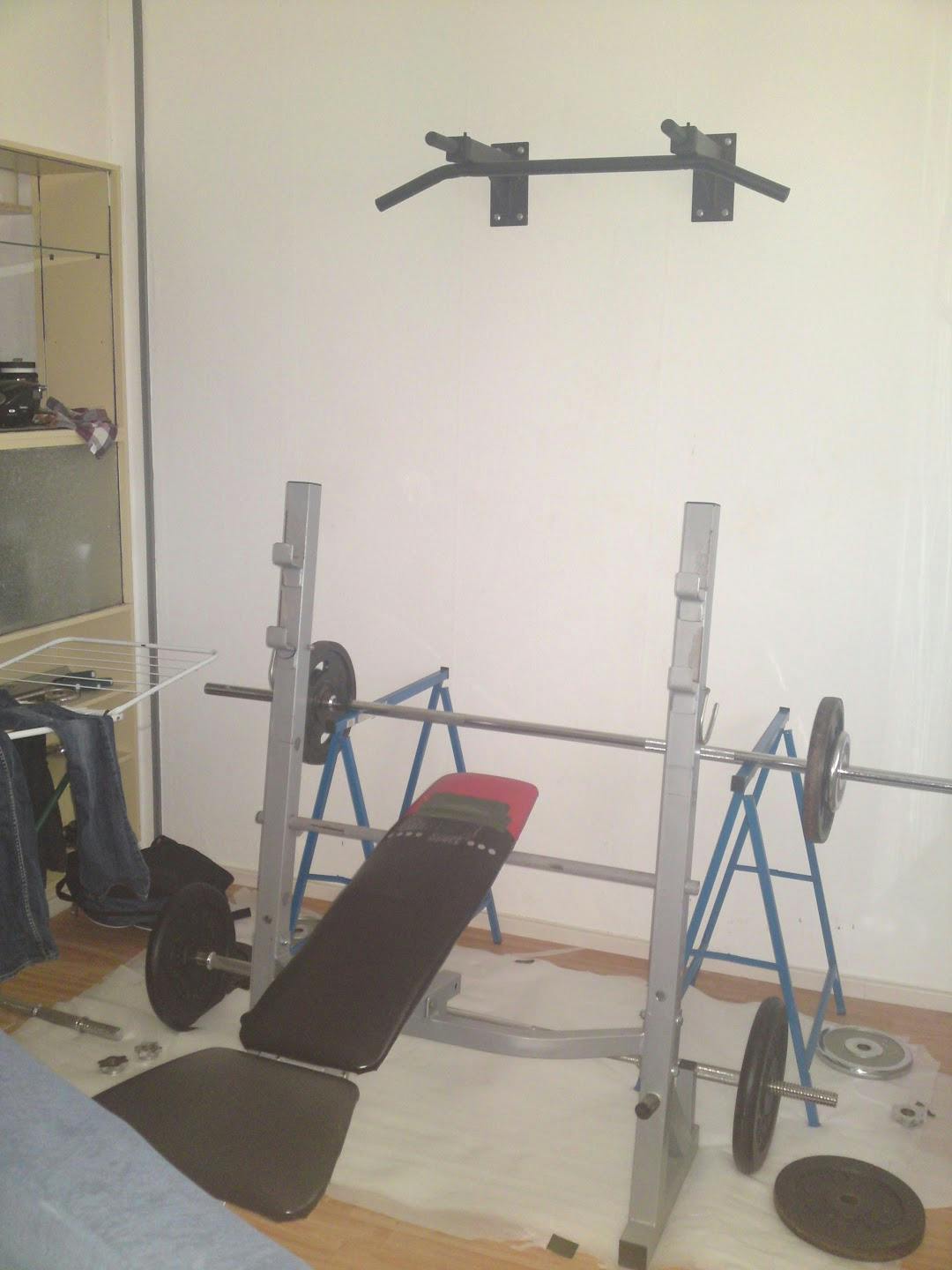 A very rudimentary home gym with a bench, barbells, weights, and a pull up bar